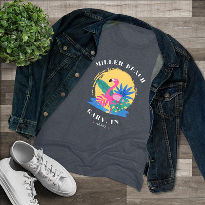 A women's triblend tee from Printify, the Miller Beach Flamingo features a blue color and "Miller Beach Gary, IN 46403" text with a vibrant design of a flamingo amidst tropical foliage for a vintage aesthetic. This t-shirt is showcased on a wooden surface alongside a denim jacket, white sneakers, and a potted plant.