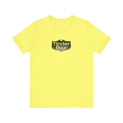 A light blue Printify "The Tinder Box 1980s" unisex jersey short sleeve tee featuring the "Tinder Box" logo in the center. The logo consists of a dark shield with the words "Tinder Box" in white text, with a small flame icon replacing the dot above the "i".