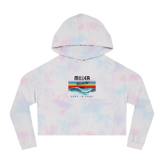 Introducing the Miller Beach Surf Women's Cropped Hooded Sweatshirt by Printify, a white personalized hoodie featuring a pastel tie-dye pattern. The front showcases a bold "Miller Beach" graphic above "Gary, IN" on a vibrant striped background with stylized mountains and waves. This long-sleeve hoodie comes in a trendy cropped style complete with a drawstring hood.