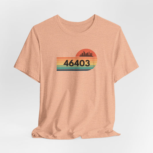 The Miller Beach 46403 Unisex Jersey Short Sleeve Tee from Printify is a peach-colored shirt featuring a retro design with "46403" in bold black text. It boasts horizontal stripes in green, yellow, and orange beneath a red-orange sunset and silhouetted city skyline.