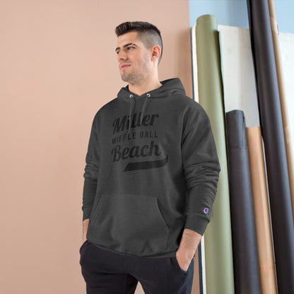 A man wearing a Champion Hoodie from Printify, featuring the "Miller Beach Wiffel Ball Retro Jersey Logo," stands against a peach-colored wall and vertical rolls of various colored fabrics. He has short, dark hair and is looking slightly to the left with a relaxed expression.