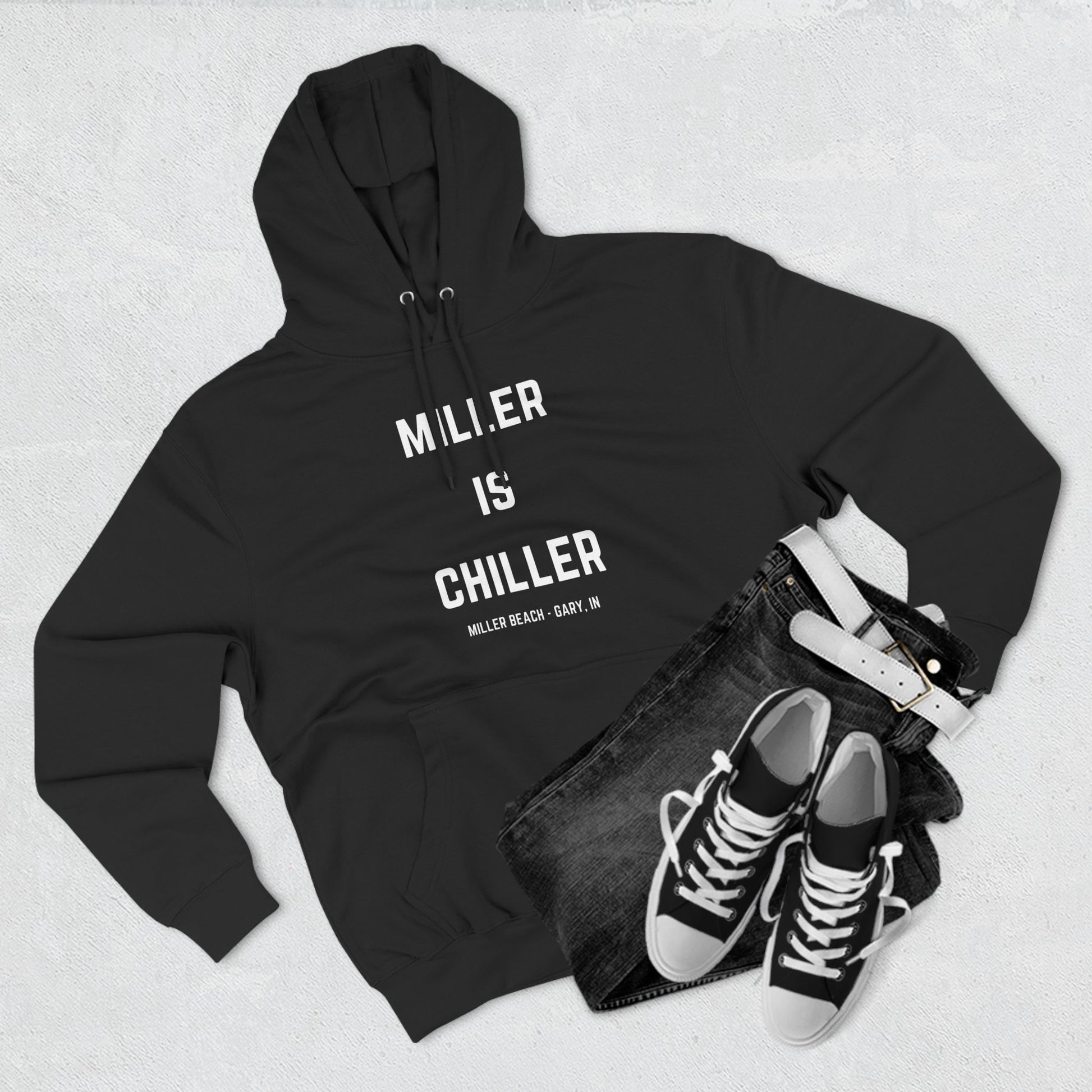 The Miller is Chiller - Miller Beach Three-Panel Fleece Hoodie from Printify features bold white text reading "MILLER IS CHILLER" on the front, with smaller text below stating "MILLER BEACH - GARY, IN." This comfortable black hoodie includes a front pocket and drawstrings.