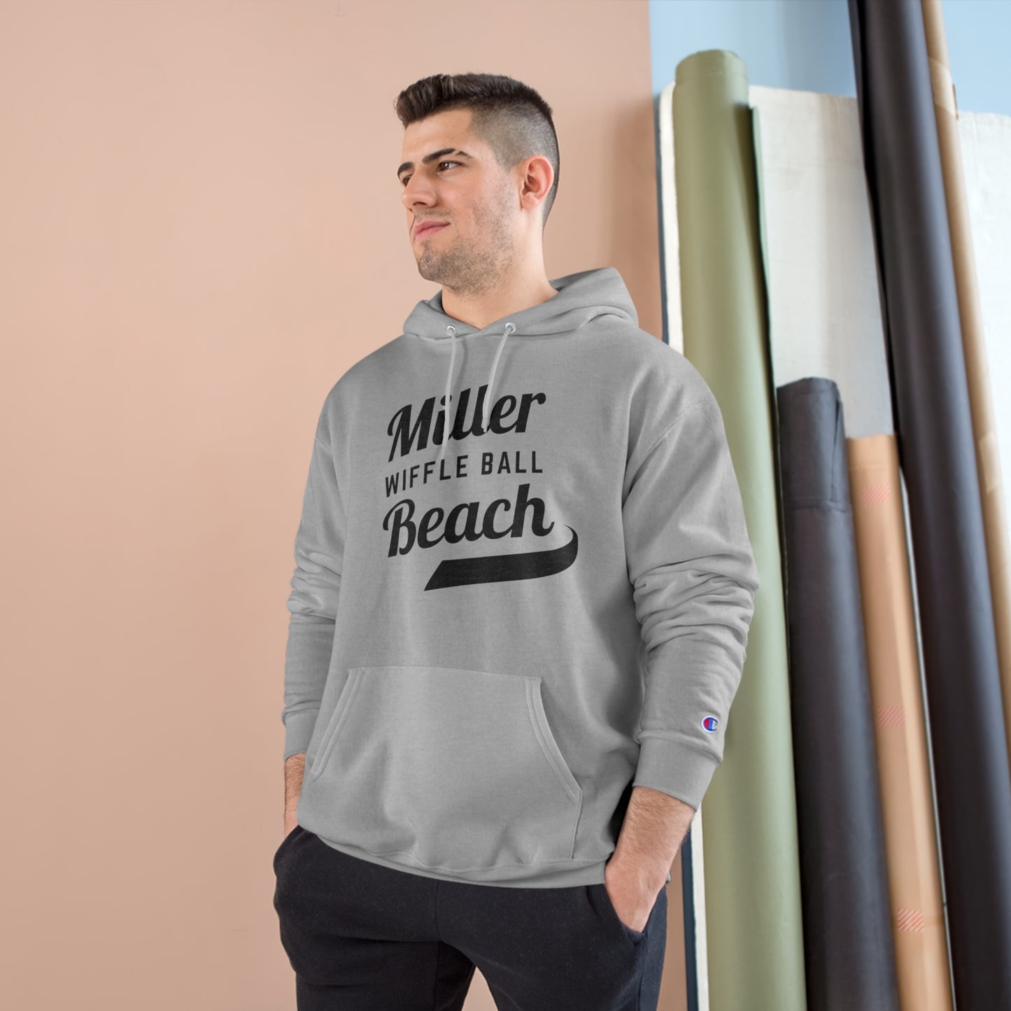 A man wearing a Champion Hoodie from Printify, featuring the "Miller Beach Wiffel Ball Retro Jersey Logo," stands against a peach-colored wall and vertical rolls of various colored fabrics. He has short, dark hair and is looking slightly to the left with a relaxed expression.