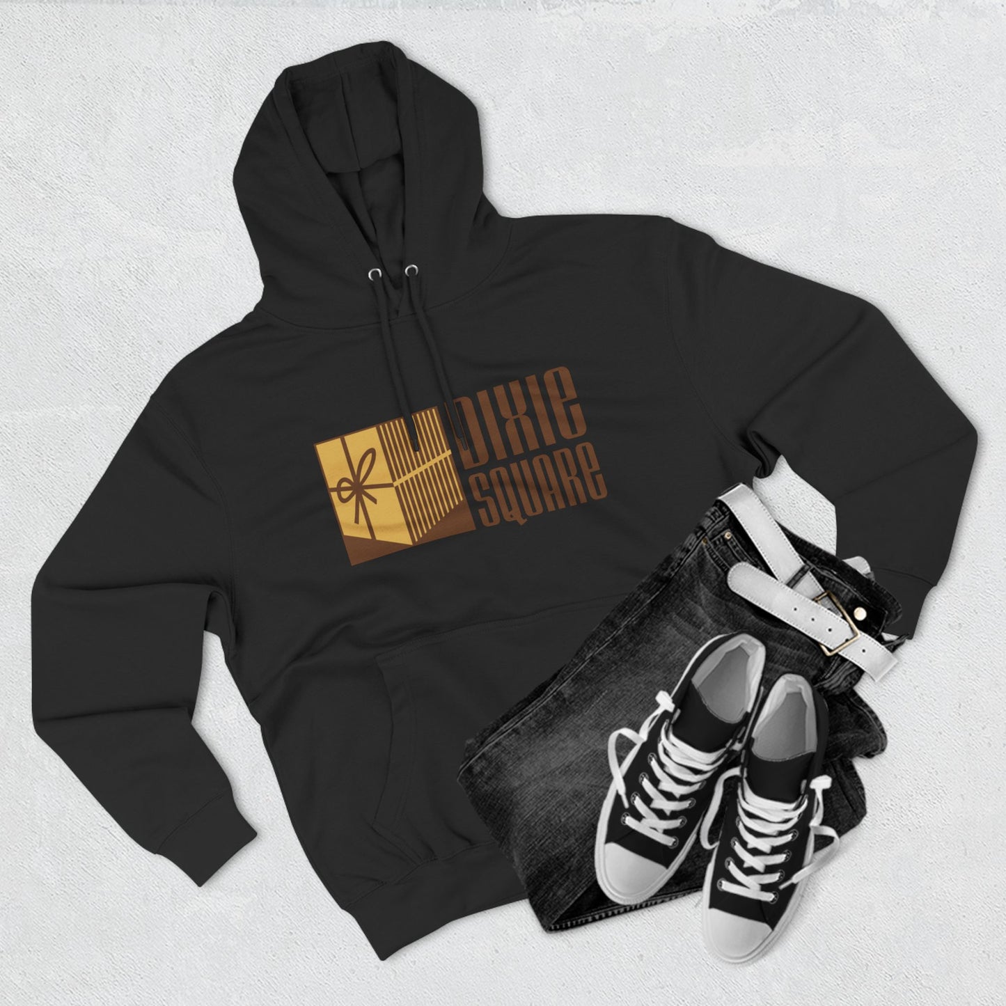 A black hoodie from Printify, named "Dixie Square 1960s Logo - Harvey, IL - Three-Panel Fleece Hoodie," featuring the words "Dixie Square" in brown stylized block letters on the front. Embracing a retro charm, the design also includes a graphic of a gold-colored square with a scissors icon and vertical lines, reminiscent of Dixie Square Mall's heyday.