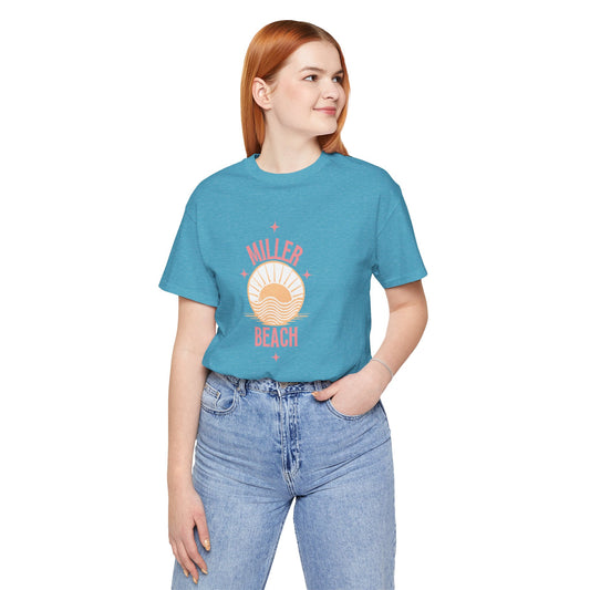 A person with red hair, wearing a Printify Miller Beach Compass unisex jersey short sleeve tee that features "Miller Beach" in pink letters alongside a sun graphic, stands with one hand in their pocket and smiles while looking to the side. The 100% Airlume combed and ring-spun cotton shirt is paired with light-wash jeans.