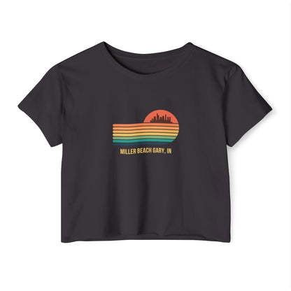 The Miller Beach Gary, IN Gradient Sunset - Women's Festival Crop Top by Printify is a mauve-colored T-shirt showcasing a retro-style graphic design of a sunset with multicolored stripes and a city skyline silhouette. Ideal for festival season, the text below reads "Miller Beach Gary, IN." Crafted to be soft and durable, this custom crop top provides both comfort and style throughout the day.