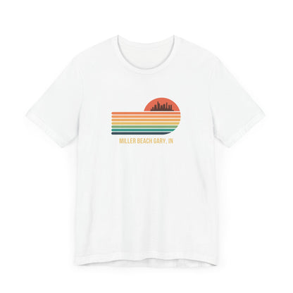 Introducing the Miller Beach Gary, IN Sunset Gradient - Unisex Jersey Short Sleeve Tee by Printify. This stylish blue T-shirt features a circular sunset graphic on the chest, showcasing a black city skyline silhouette against an orange-red sunset with horizontal stripes in green, yellow, and orange. Below the graphic reads "MILLER BEACH GARY, IN." Crafted from soft cotton for extra beachy charm.