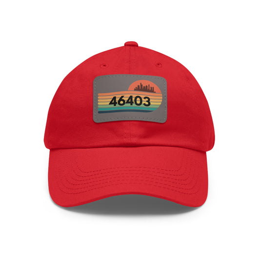 The Miller Beach Dad Hat from Printify, in a striking red and crafted from bio-washed chino twill, showcases a rectangular leather patch on the front. The patch features a stylized silhouette of a city skyline in black atop horizontal stripes in green, yellow, and orange, with the number "46403" prominently printed in bold black text.