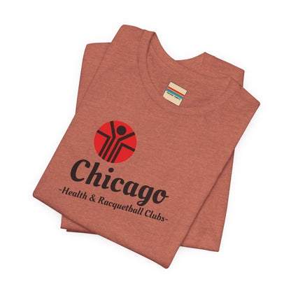 Two gray T-shirts are neatly folded on top of each other, with the top shirt showcasing the text "Chicago Health & Racquetball Clubs" in black below a red logo featuring a person with raised arms inside a circular design. This retro-inspired tee, named "Chicago Health Clubs 1980s Retro - Unisex Jersey Short Sleeve Tee" by Printify, is perfect for any fan of Chicago Health Clubs.