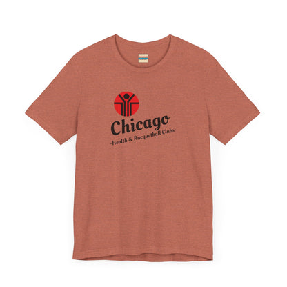 Two gray T-shirts are neatly folded on top of each other, with the top shirt showcasing the text "Chicago Health & Racquetball Clubs" in black below a red logo featuring a person with raised arms inside a circular design. This retro-inspired tee, named "Chicago Health Clubs 1980s Retro - Unisex Jersey Short Sleeve Tee" by Printify, is perfect for any fan of Chicago Health Clubs.