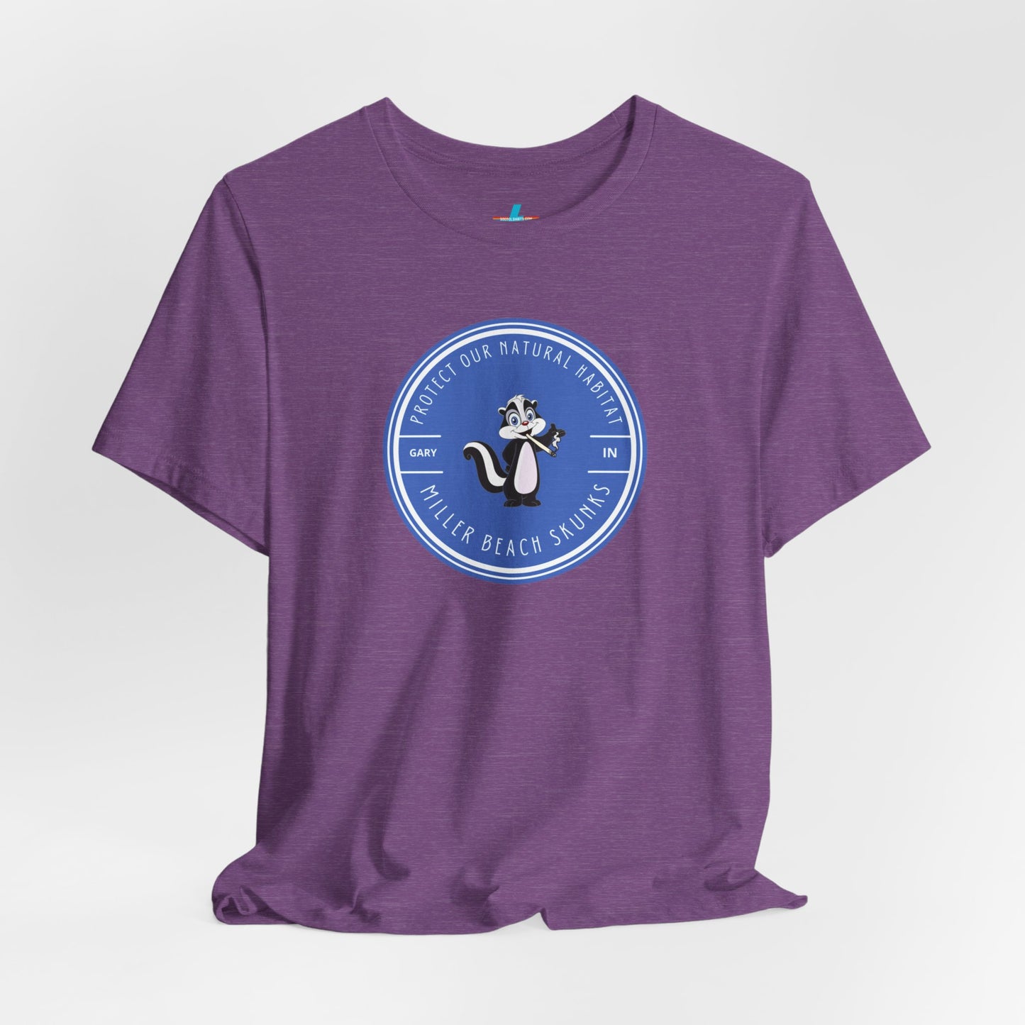 The Printify Miller Beach Skunks - Unisex Jersey Short Sleeve Tee is a gray T-shirt that features a circular blue logo at the center. Inside the logo, there is an illustration of a skunk with the text "Protect Our Habitat" and "Miller Beach Skunks" around the border, promoting environmental consciousness. The upper left part of the logo reads "Gary, IN.