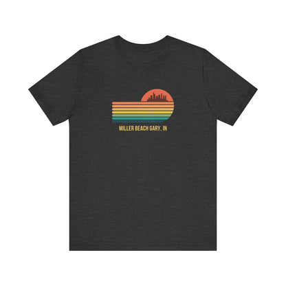 Introducing the Miller Beach Gary, IN Sunset Gradient - Unisex Jersey Short Sleeve Tee by Printify. This stylish blue T-shirt features a circular sunset graphic on the chest, showcasing a black city skyline silhouette against an orange-red sunset with horizontal stripes in green, yellow, and orange. Below the graphic reads "MILLER BEACH GARY, IN." Crafted from soft cotton for extra beachy charm.