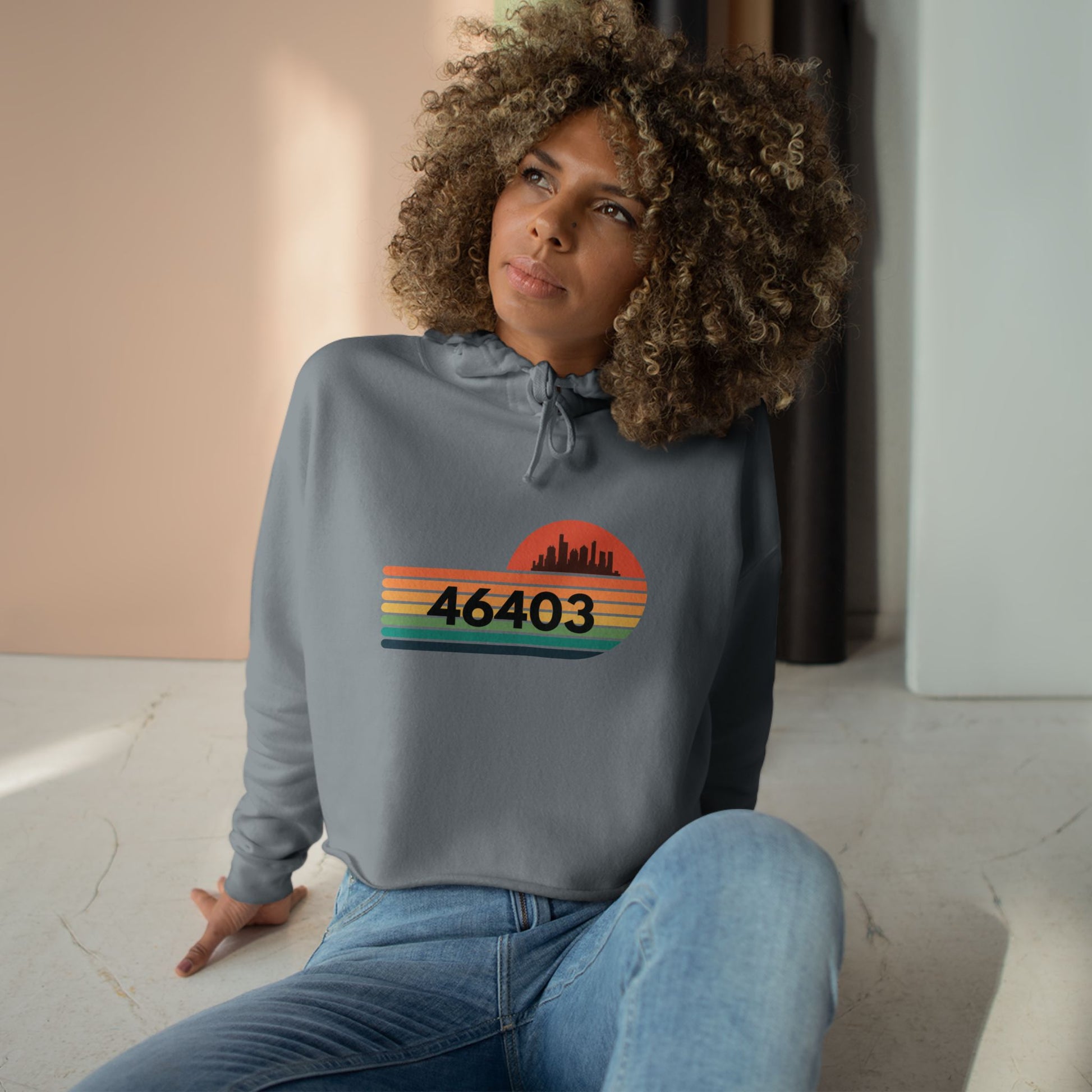 The Miller Beach 46403 Women's Crop Hoodie by Printify features a nostalgic design with a city skyline silhouette and the iconic "46403" zip code, set against a striped sunset backdrop in vibrant orange, green, and yellow hues that capture the essence of Miller Beach.