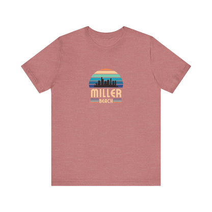 A brown unisex jersey short sleeve tee called "Miller Beach Chicago Skyline" from Printify features a vintage-style design with a sunset, the Chicago skyline, and "Miller Beach" in bold letters. The design incorporates shades of blue, orange, and yellow and is displayed against a plain white background.