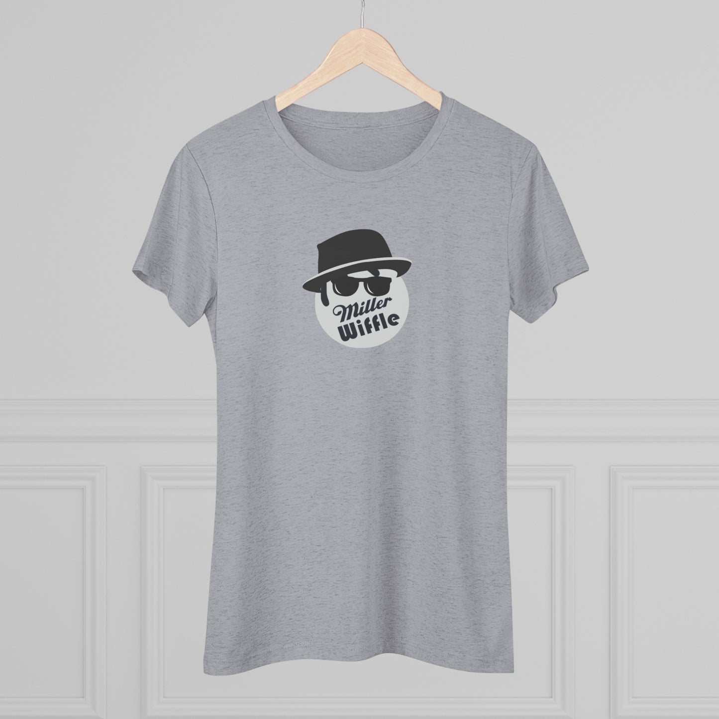 A cozy red t-shirt, the Miller Wiffel Ball - Blues Brothers - Women's Triblend Tee by Printify, features a graphic of a masked face wearing a fedora hat and sunglasses, along with the text "Miller Wiffle." The t-shirt is displayed on a wooden floor, paired with a denim jacket, white sneakers, and a potted green plant to create a vintage look.