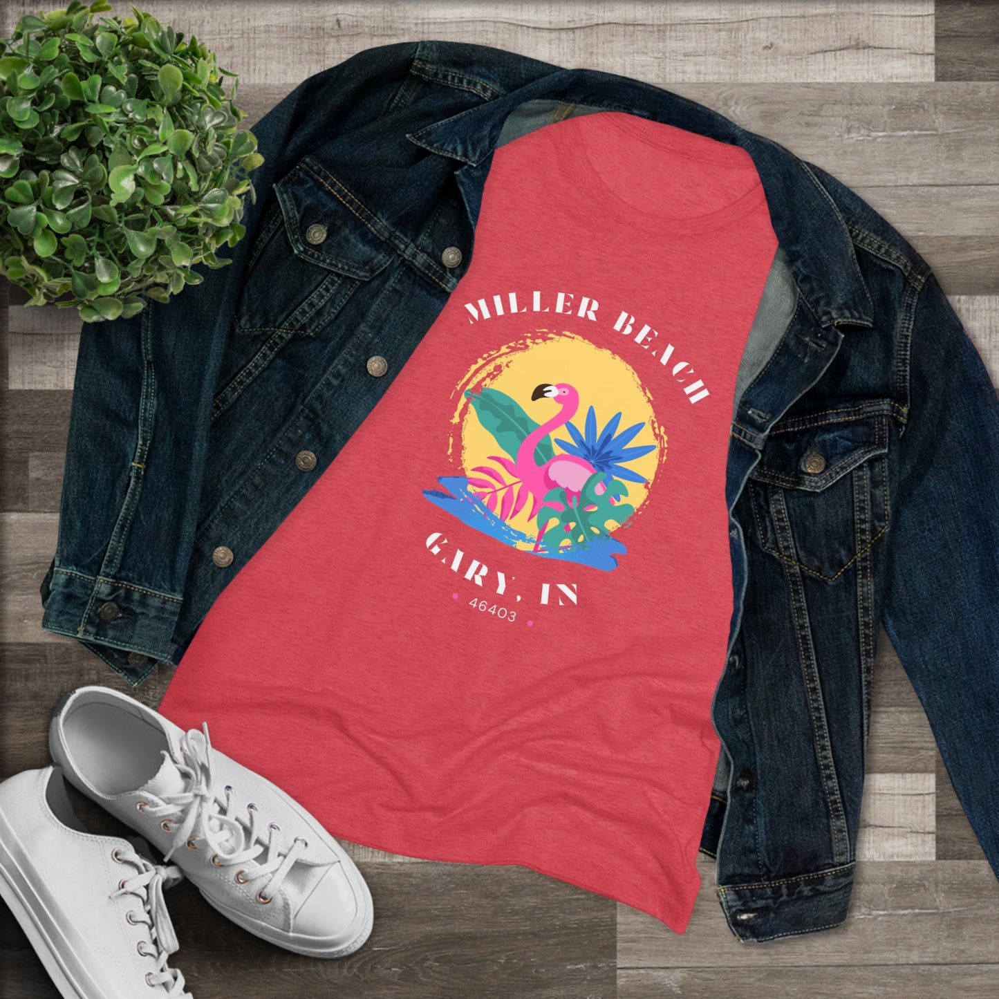 A women's triblend tee from Printify, the Miller Beach Flamingo features a blue color and "Miller Beach Gary, IN 46403" text with a vibrant design of a flamingo amidst tropical foliage for a vintage aesthetic. This t-shirt is showcased on a wooden surface alongside a denim jacket, white sneakers, and a potted plant.