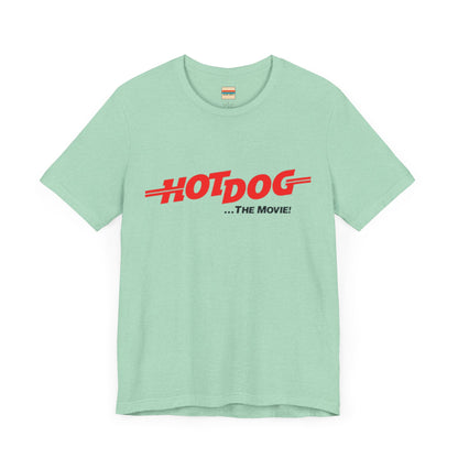 A smiling man and woman interact while wearing matching yellow "Hot Dog The Movie 1984 - Unisex Jersey Short Sleeve Tee" by Printify, featuring bold red "HOT DOG THE MOVIE" text. Their vibrant tees perfectly complement their blue jeans, with the man casually having his hands in his pockets and the woman resting one hand on his shoulder.