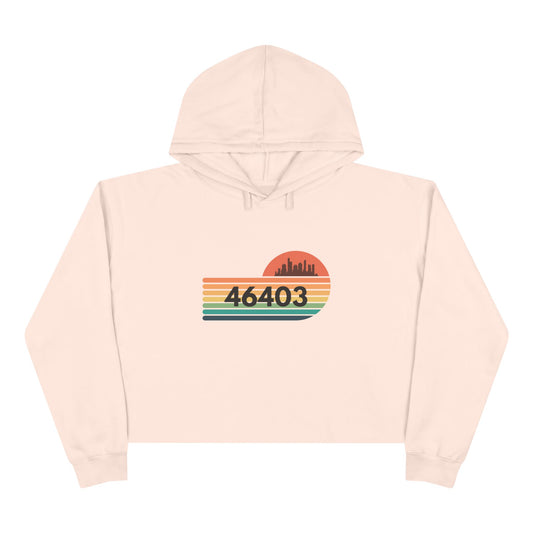 The Miller Beach 46403 Women's Crop Hoodie by Printify features a nostalgic design with a city skyline silhouette and the iconic "46403" zip code, set against a striped sunset backdrop in vibrant orange, green, and yellow hues that capture the essence of Miller Beach.