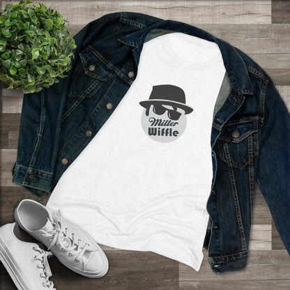 A cozy red t-shirt, the Miller Wiffel Ball - Blues Brothers - Women's Triblend Tee by Printify, features a graphic of a masked face wearing a fedora hat and sunglasses, along with the text "Miller Wiffle." The t-shirt is displayed on a wooden floor, paired with a denim jacket, white sneakers, and a potted green plant to create a vintage look.