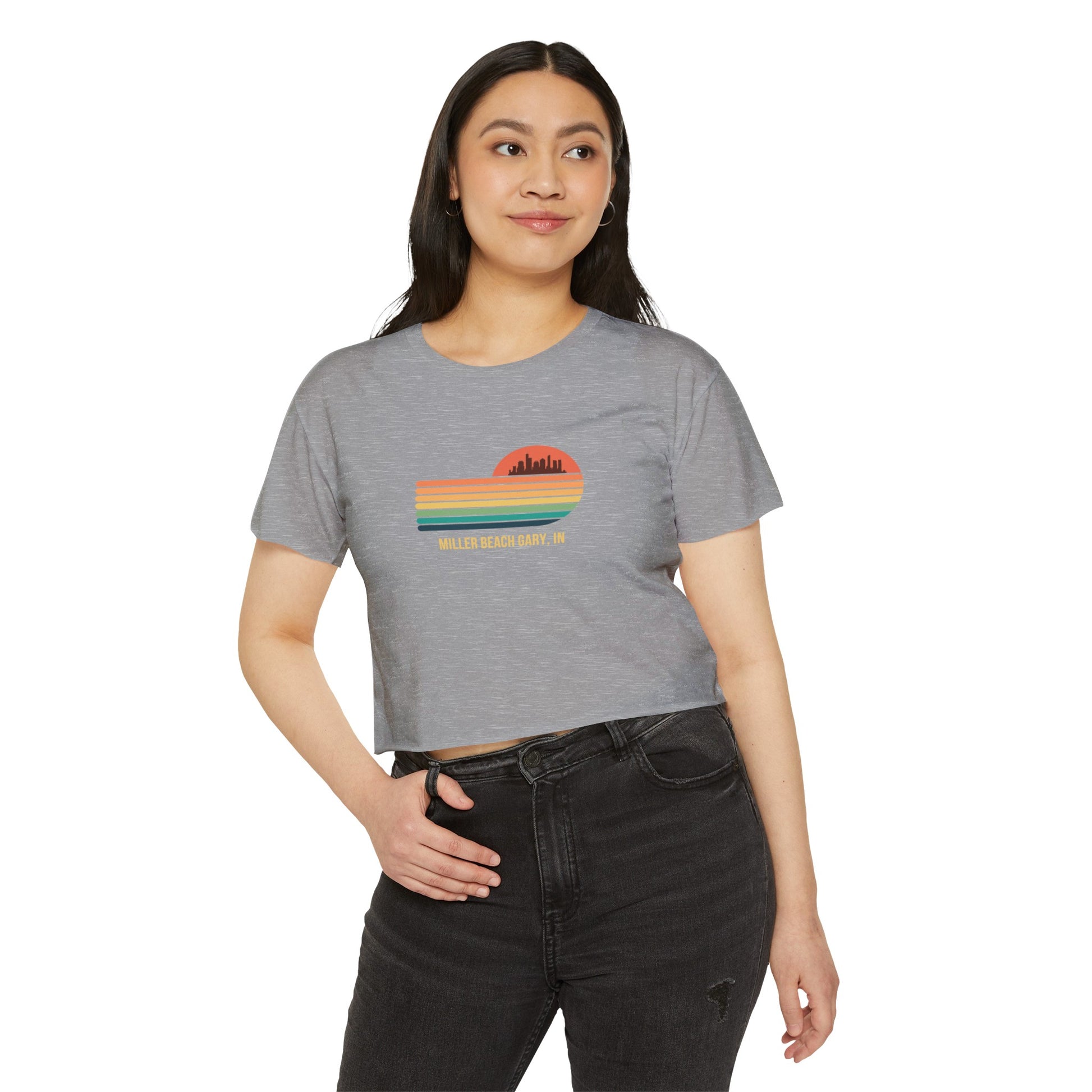 The Miller Beach Gary, IN Gradient Sunset - Women's Festival Crop Top by Printify is a mauve-colored T-shirt showcasing a retro-style graphic design of a sunset with multicolored stripes and a city skyline silhouette. Ideal for festival season, the text below reads "Miller Beach Gary, IN." Crafted to be soft and durable, this custom crop top provides both comfort and style throughout the day.