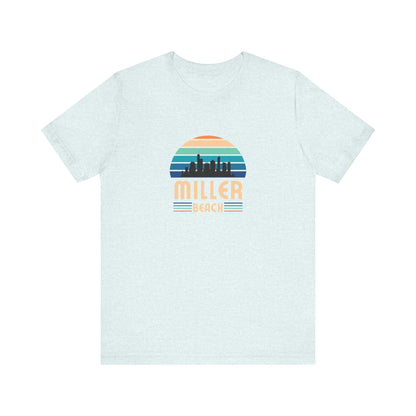 The Printify Miller Beach Chicago Skyline - Unisex Jersey Short Sleeve Tee is a light blue T-shirt featuring a stylized graphic of the Chicago skyline against a setting sun with gradient shades of blue and orange. Below the graphic, "Miller Beach" is printed in bold, yellow letters.