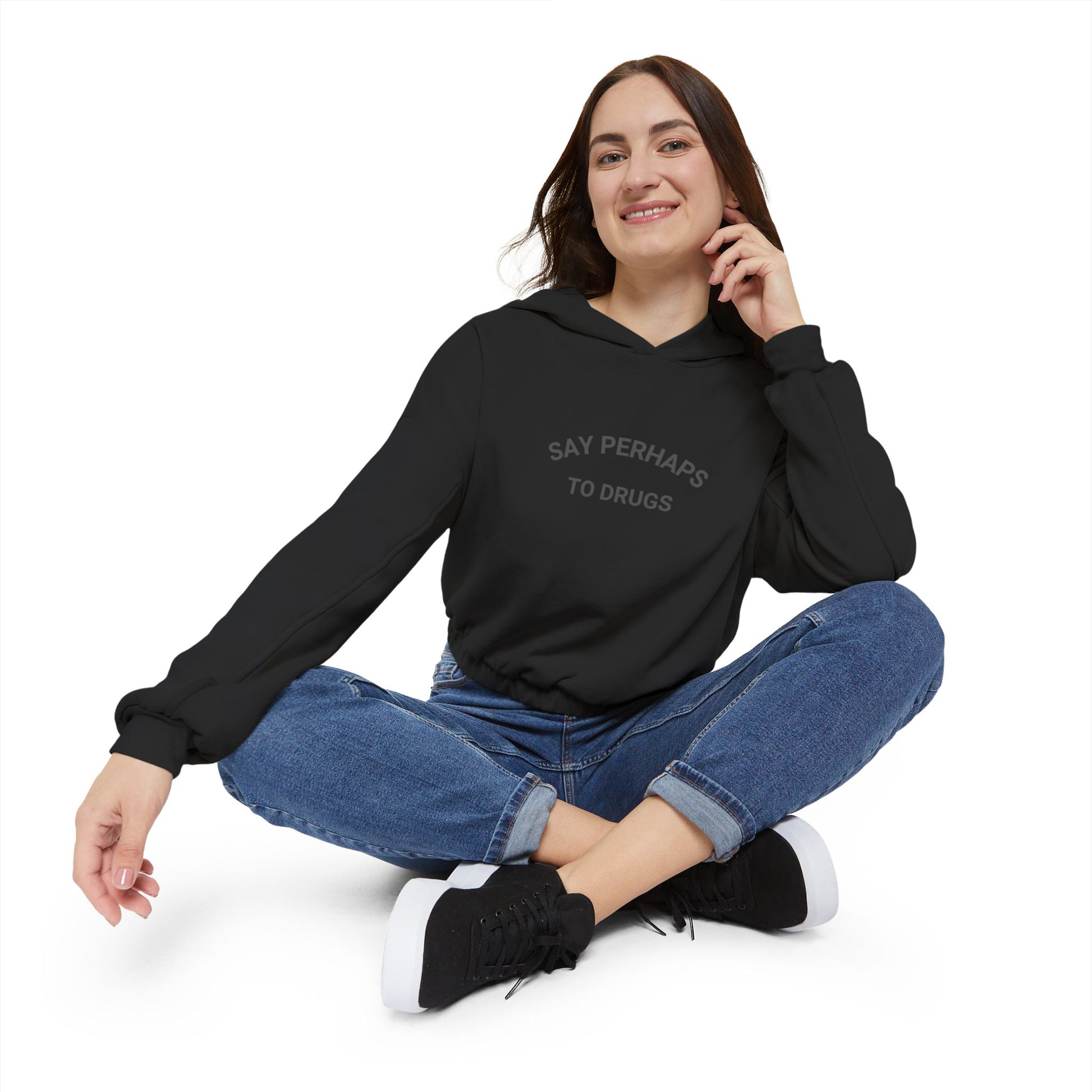 A person confidently stands in front of the camera with one hand on their hip, wearing a Printify "Say Perhaps to Drugs" Women's Cinched Bottom Hoodie. Crafted from soft Airlume cotton, this light-colored cropped hoodie pairs stylishly with blue jeans.