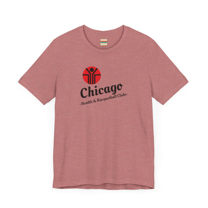 Two gray T-shirts are neatly folded on top of each other, with the top shirt showcasing the text "Chicago Health & Racquetball Clubs" in black below a red logo featuring a person with raised arms inside a circular design. This retro-inspired tee, named "Chicago Health Clubs 1980s Retro - Unisex Jersey Short Sleeve Tee" by Printify, is perfect for any fan of Chicago Health Clubs.