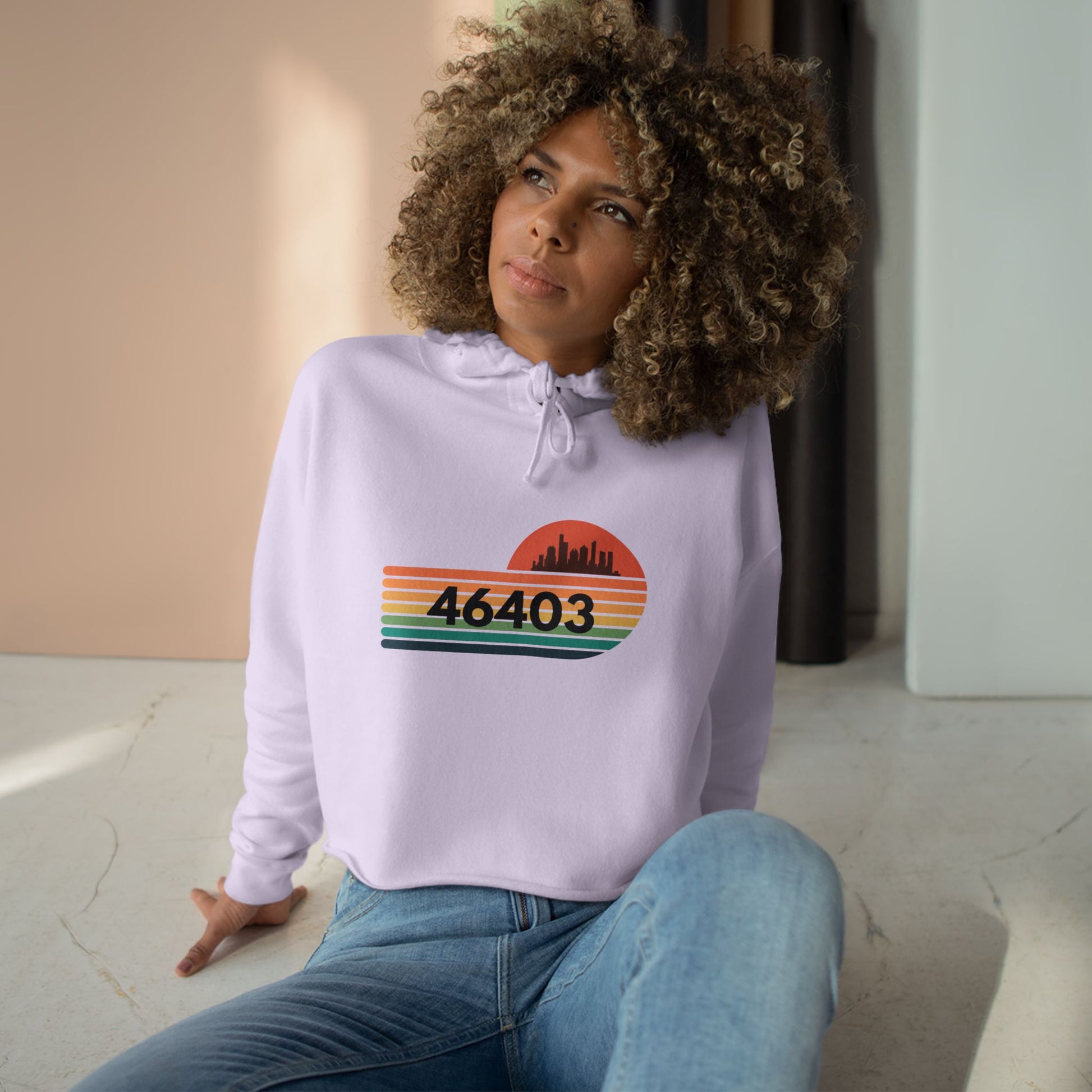 The Miller Beach 46403 Women's Crop Hoodie by Printify features a nostalgic design with a city skyline silhouette and the iconic "46403" zip code, set against a striped sunset backdrop in vibrant orange, green, and yellow hues that capture the essence of Miller Beach.