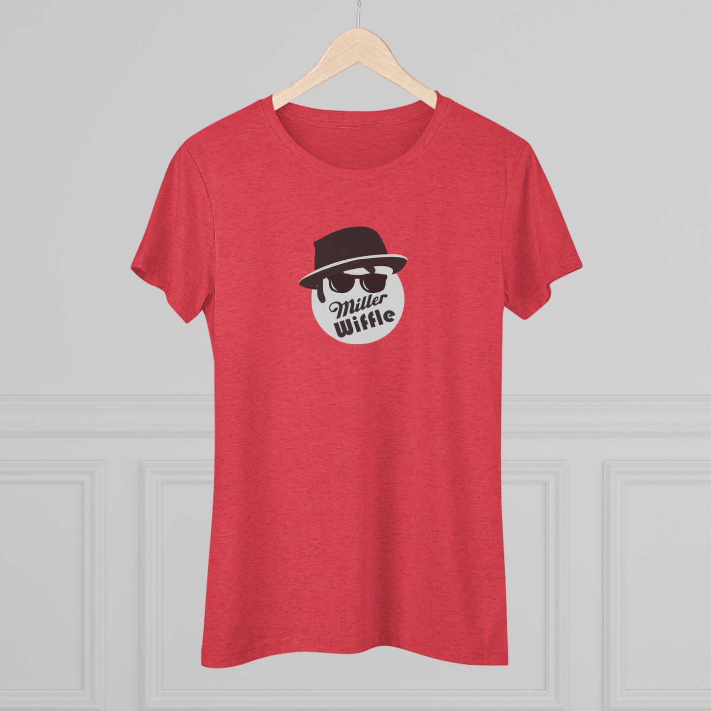 A cozy red t-shirt, the Miller Wiffel Ball - Blues Brothers - Women's Triblend Tee by Printify, features a graphic of a masked face wearing a fedora hat and sunglasses, along with the text "Miller Wiffle." The t-shirt is displayed on a wooden floor, paired with a denim jacket, white sneakers, and a potted green plant to create a vintage look.