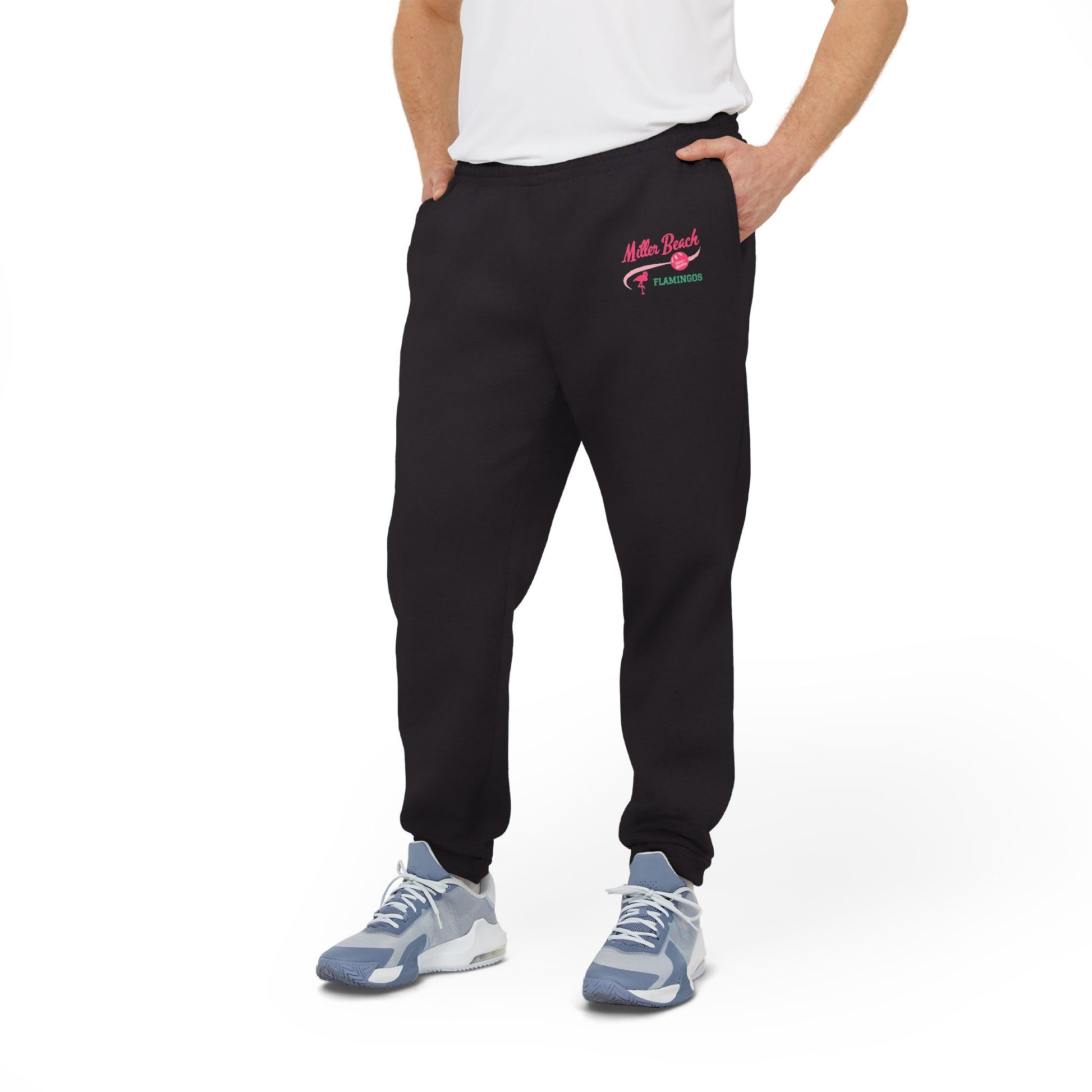 A person is confidently dressed in the cozy Miller Beach Flamingos - Adidas Unisex Fleece Joggers from Printify, featuring "Myrtle Beach" and "Yearround" text in pink and green on the left thigh. They complete their look with a white shirt and gray sneakers, standing against a plain white background.