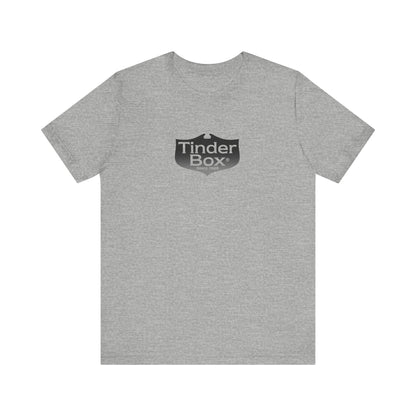 A light blue Printify "The Tinder Box 1980s" unisex jersey short sleeve tee featuring the "Tinder Box" logo in the center. The logo consists of a dark shield with the words "Tinder Box" in white text, with a small flame icon replacing the dot above the "i".