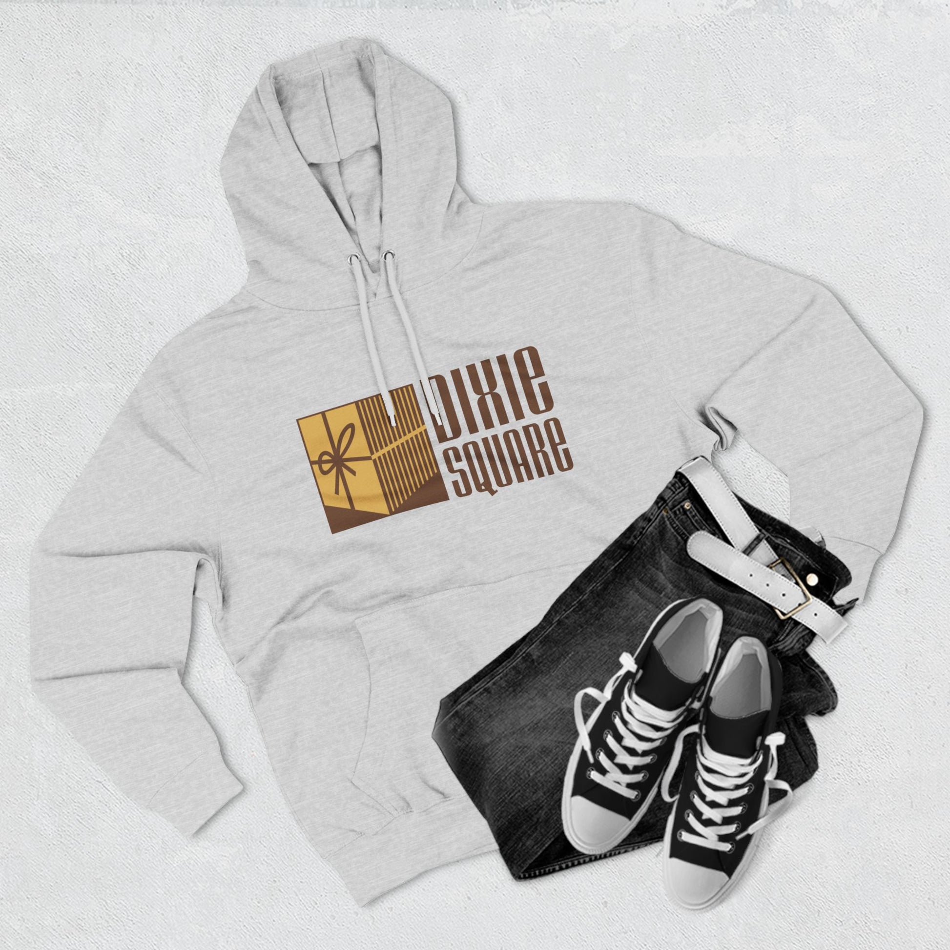 A black hoodie from Printify, named "Dixie Square 1960s Logo - Harvey, IL - Three-Panel Fleece Hoodie," featuring the words "Dixie Square" in brown stylized block letters on the front. Embracing a retro charm, the design also includes a graphic of a gold-colored square with a scissors icon and vertical lines, reminiscent of Dixie Square Mall's heyday.