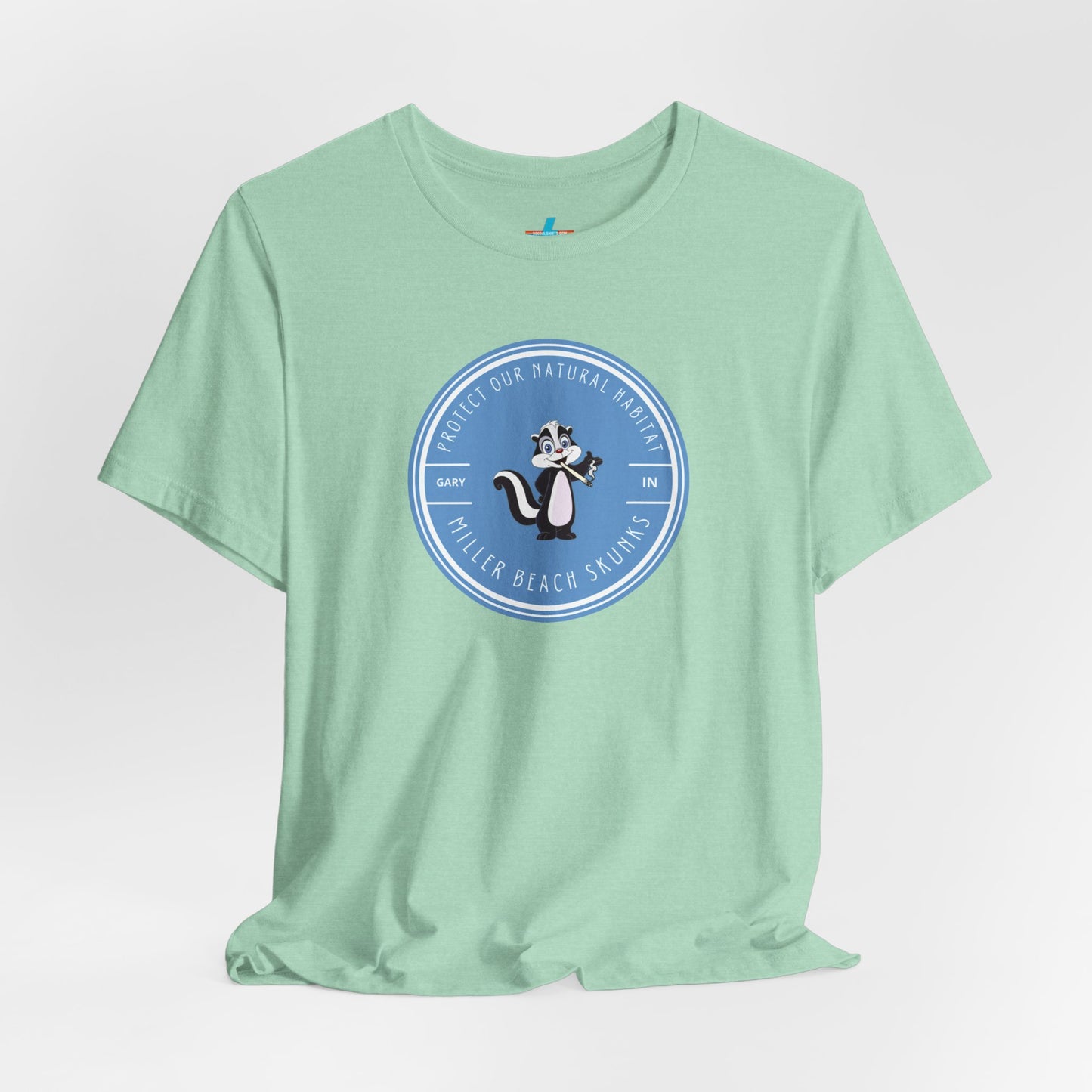 The Printify Miller Beach Skunks - Unisex Jersey Short Sleeve Tee is a gray T-shirt that features a circular blue logo at the center. Inside the logo, there is an illustration of a skunk with the text "Protect Our Habitat" and "Miller Beach Skunks" around the border, promoting environmental consciousness. The upper left part of the logo reads "Gary, IN.