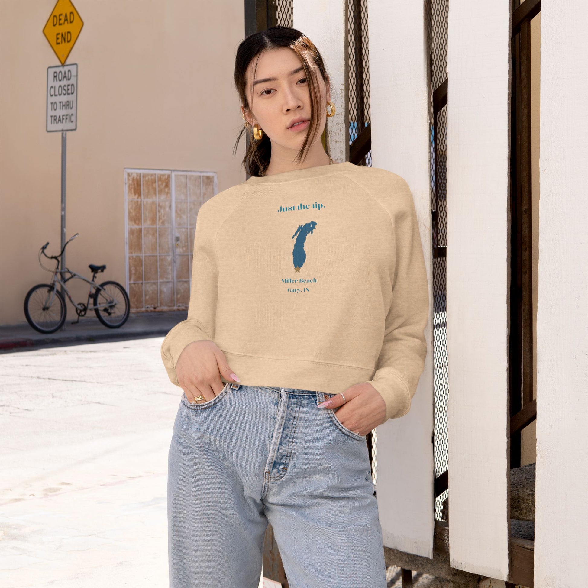 The "Just the Tip - Miller Beach" Women's Cropped Fleece Pullover by Printify is a beige sweatshirt with long sleeves and a crew neck. It features a blue-green abstract feather design in the center with the words "Just the tip" above it, and "Silly Peach" along with "Gary, IN." below. This pullover is crafted from premium tri-blend fabric for ultimate comfort and style.