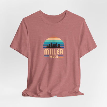 A brown unisex jersey short sleeve tee called "Miller Beach Chicago Skyline" from Printify features a vintage-style design with a sunset, the Chicago skyline, and "Miller Beach" in bold letters. The design incorporates shades of blue, orange, and yellow and is displayed against a plain white background.