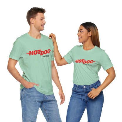 A smiling man and woman interact while wearing matching yellow "Hot Dog The Movie 1984 - Unisex Jersey Short Sleeve Tee" by Printify, featuring bold red "HOT DOG THE MOVIE" text. Their vibrant tees perfectly complement their blue jeans, with the man casually having his hands in his pockets and the woman resting one hand on his shoulder.