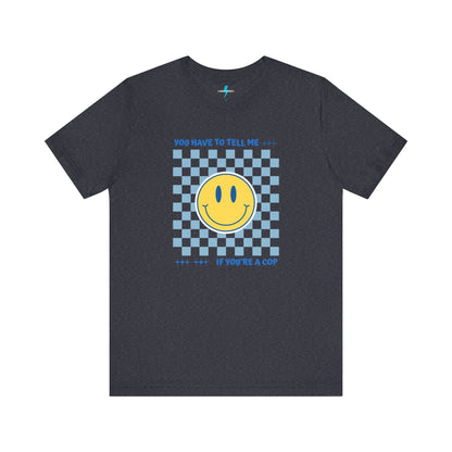 A light yellow unisex jersey short sleeve tee from Printify featuring a blue and yellow checkerboard pattern with a large yellow smiley face in the center. The shirt displays blue text above and below the pattern that reads, "You have to tell me if you're a cop." It's made from 100% Airlume combed and ring-spun cotton.