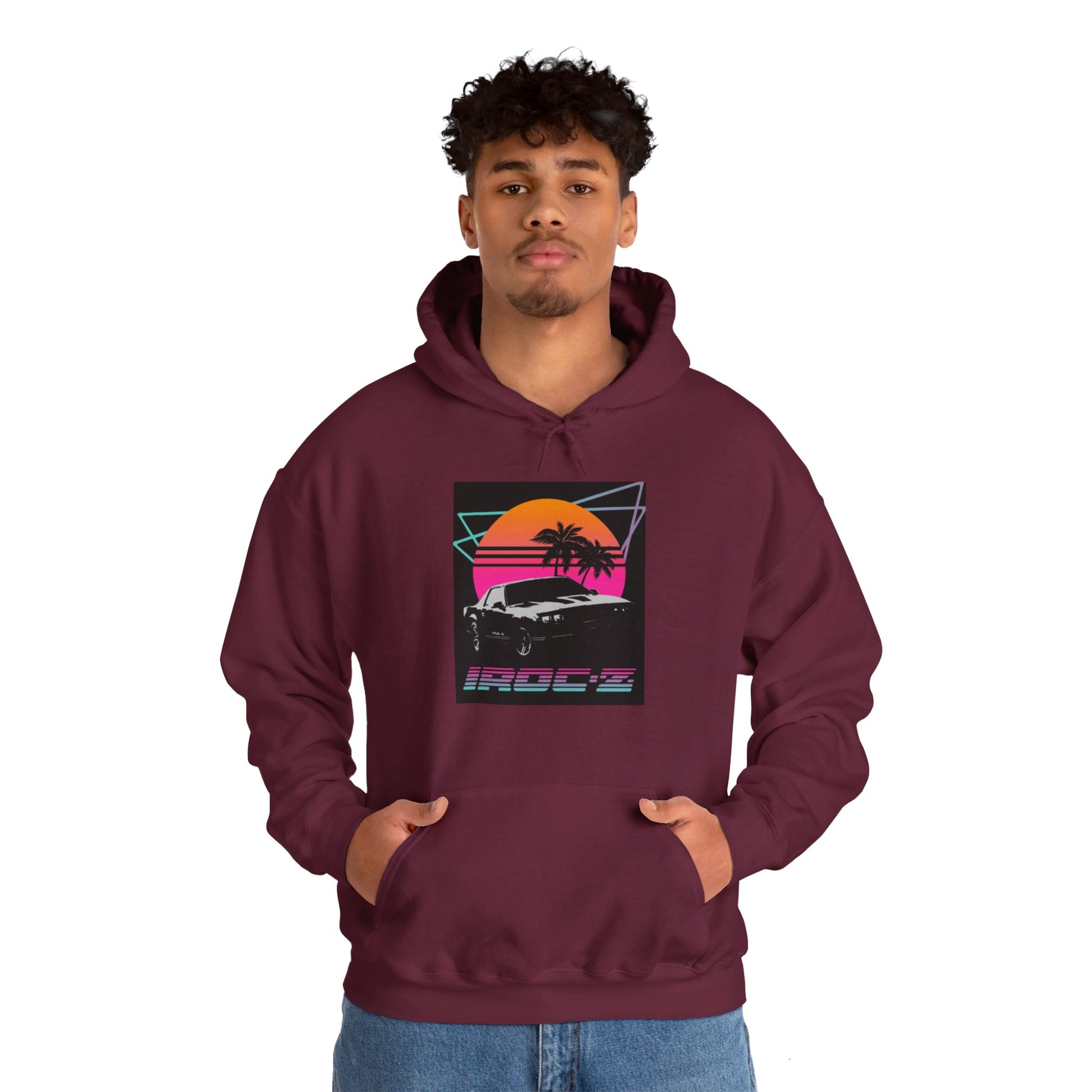 Introducing the Chevrolet IRoc Z28 - 1980s Retro Hoodie by Printify: This vibrant pink hoodie showcases a striking retro design on the front, featuring a classic American muscle car set against an orange and red sunset with palm trees, intersected by geometric shapes. The text "IROCZ" is prominently displayed below the image. Offering a relaxed fit and equipped with a front pocket, this hoodie perfectly captures the essence of 1980s style.